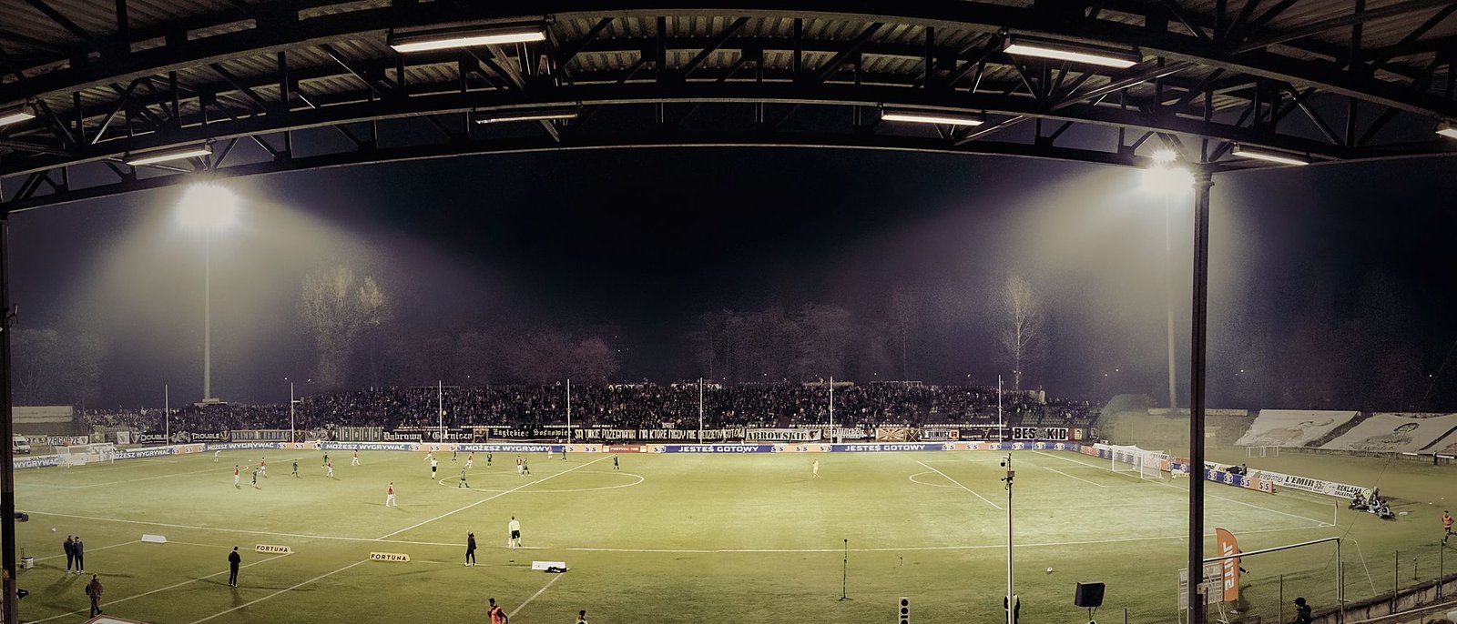 A football stadium at night under floodlights, players on the illuminated pitch and a full crowd in the stands